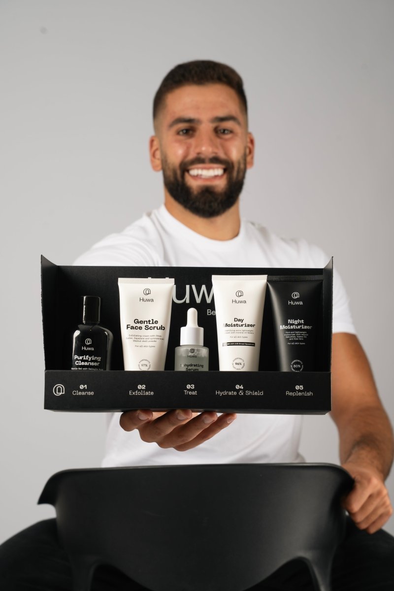 Smiling and confident Man holding a black box with skincare products on a gray background it is the huwa complete skincare bundle