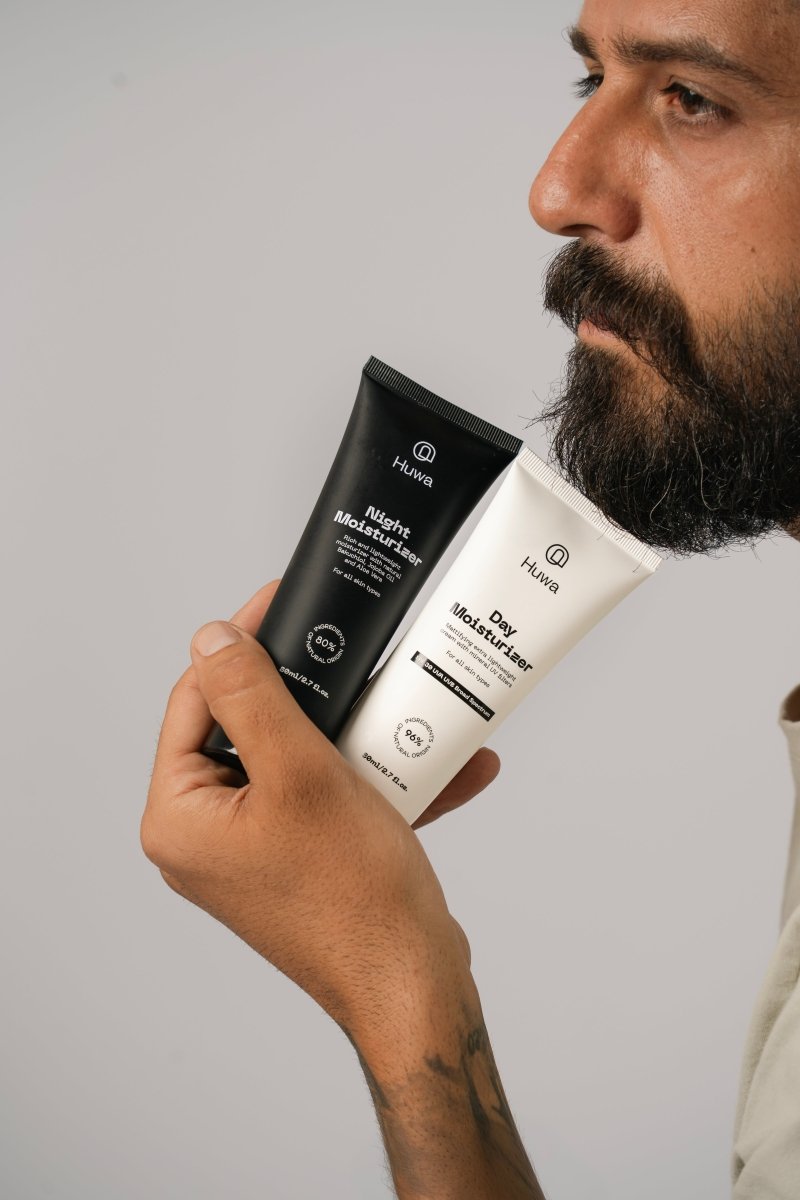 Man holding the huwa skincare night moisturizer and day moisturizer next to his shin with his groomed beard and clear skin and holding both tubes with same hand on a neutral background