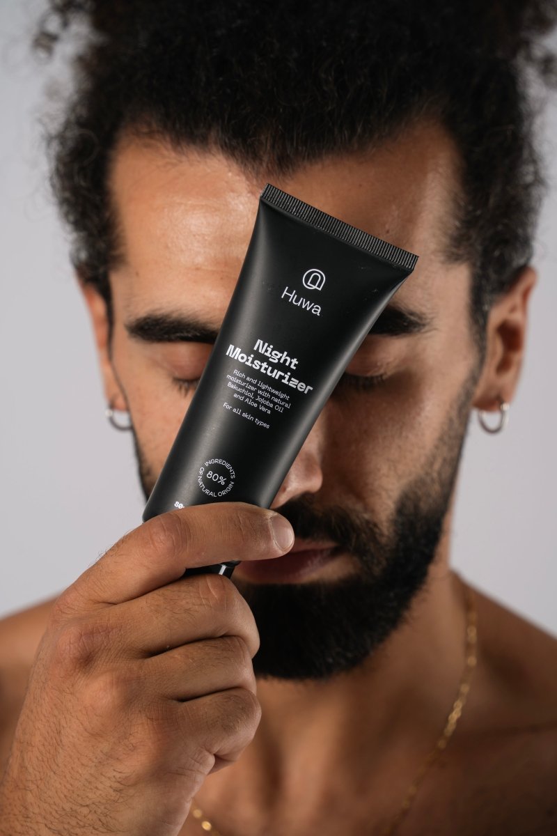 Man holding a black skincare tube labeled Huwa skincare close to his face with closed eyes and curly hair and clear skin against a neutral background