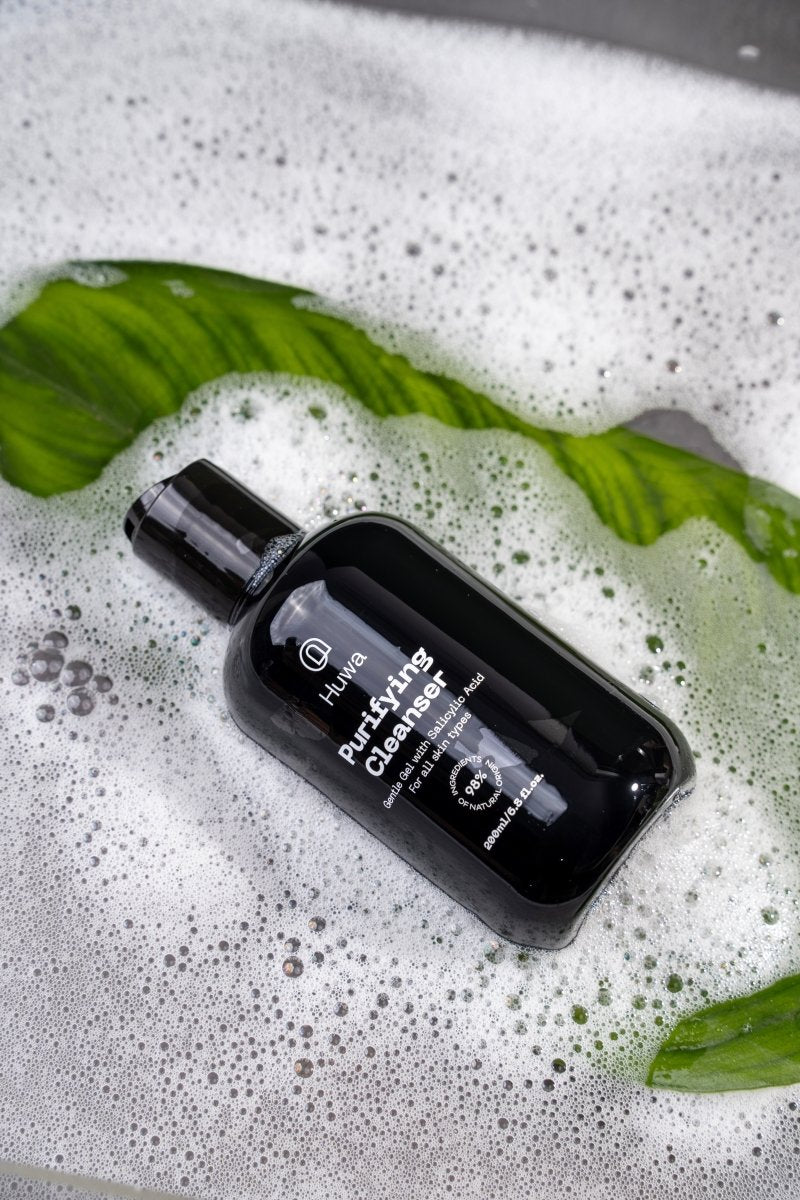 black bottle of purifying cleanser of huwa skincare in soap and water and a leaf under it