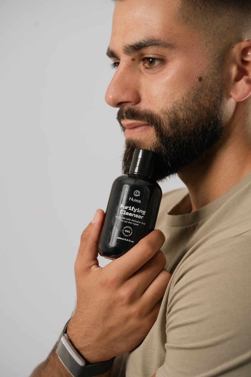 The Huwa Skincare Cleanser a purifying cleanser with salicylic acid suitable for all skin types held next to chin of the man with a beard with a gray background and the man wearing a whoop