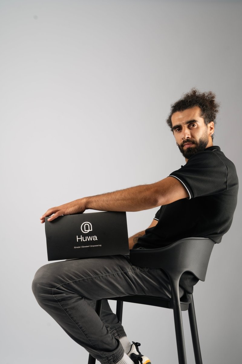 Man holding a Set of skincare products including a scrub a cleanser and day moisturizer and night moisturizer and a serum sitting on a chair and looking sharp with curly hair showing the bundle