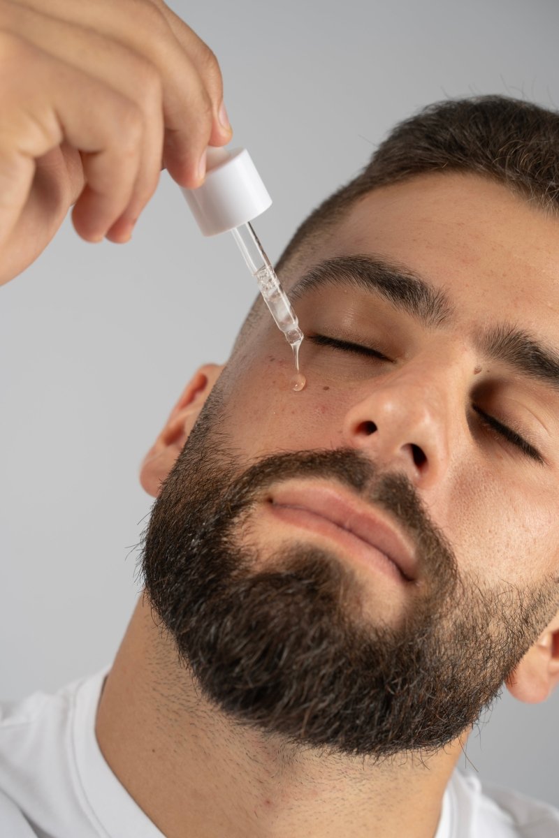 Man applying huwa skincare serum from a white dropper of liquid to his face against a neutral background while closing his eyes and full of hydration