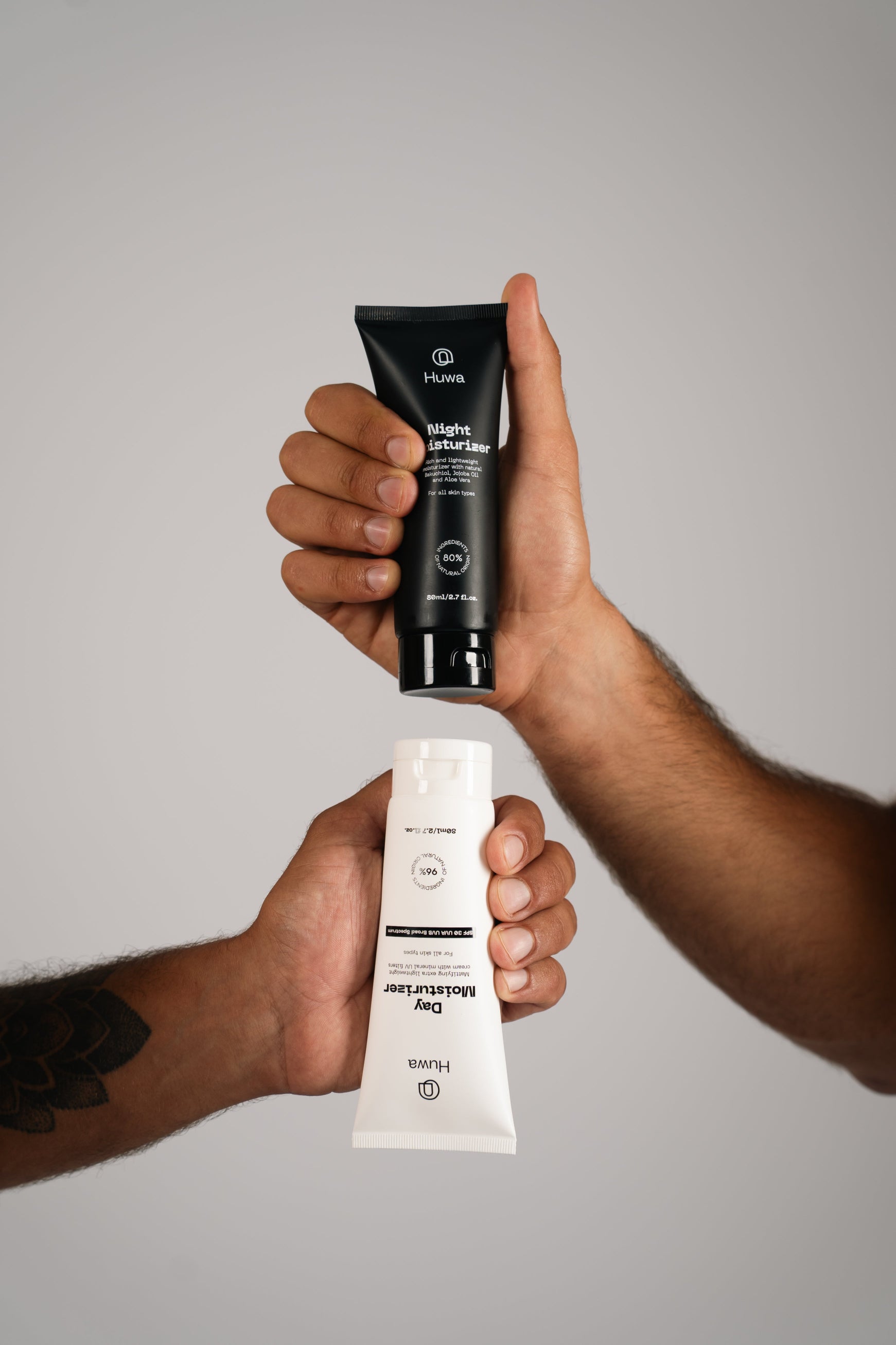Two hands holding a black and white skincare product tube against a neural background one is the day moisturizer and the other is night moisturizer of huwa skincare 